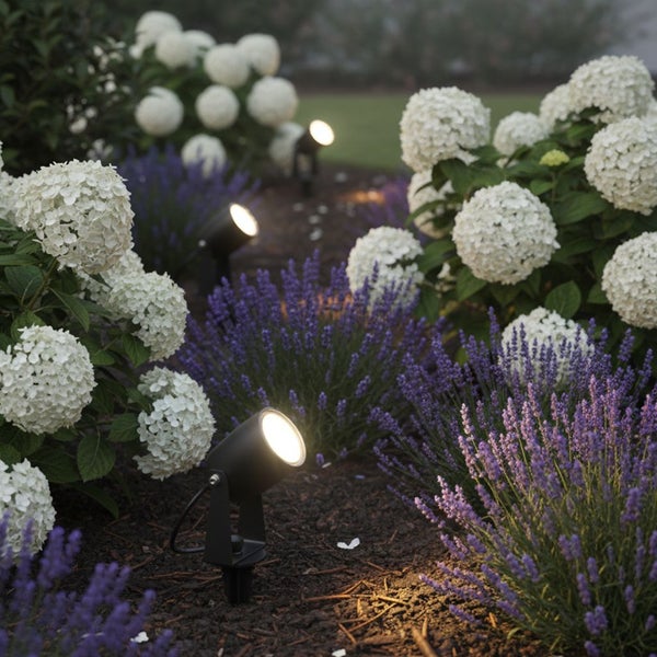 LED-Gartenstrahler mit Erdspieß beleuchten weiße Hortensien und violetten Lavendel in einem Gartenbeet bei Dämmerung.