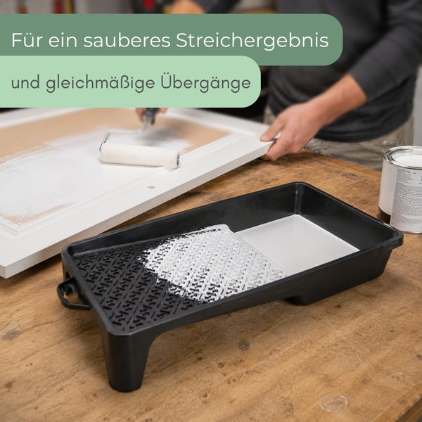 Eine Person streicht eine weiße Fläche mit einer Farbwalze, im Vordergrund steht eine schwarze Farbwanne mit weißer Farbe auf einem Holztisch.