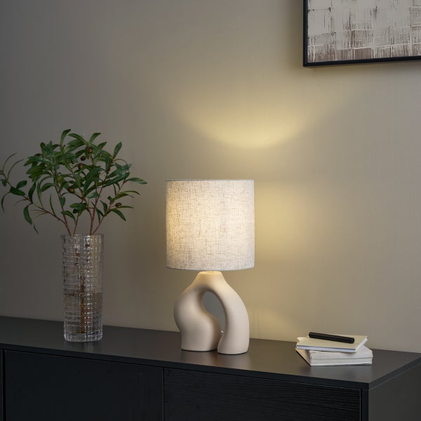 Moderne Tischleuchte mit organisch geformtem Keramikfuß und Leinenschirm auf einem dunklen Sideboard neben einer Vase mit Zweigen.