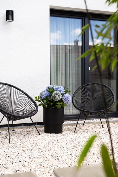 Moderne Terrasse mit zwei schwarzen runden Gartenstühlen, einem schwarzen Pflanzkübel mit Hortensien auf weißem Kies vor einer Hauswand.