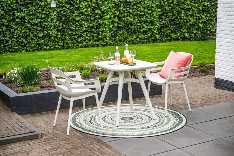 Modernes Gartenmöbel-Set mit weißem quadratischem Tisch und zwei Armlehnenstühlen auf einem gemusterten Outdoor-Teppich auf einer Terrasse.
