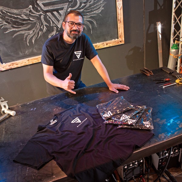 Schwarzes T-Shirt mit Stahlwerk-Logo auf einem Tisch, mit einer Person, die einen Daumen nach oben zeigt.