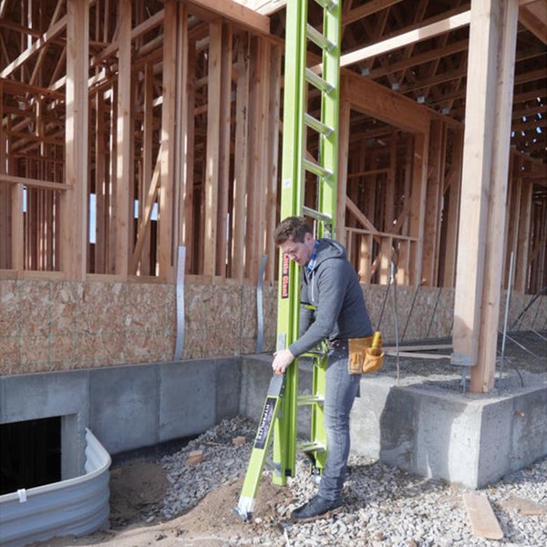 Ein Mann stellt eine grüne Little Giant Schiebeleiter an einer Holzrahmenkonstruktion auf einer Baustelle auf.