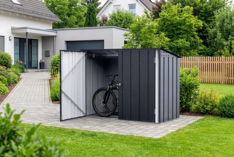 Dunkelgraue Fahrradbox aus Metall mit geöffneter Tür und einem Mountainbike im Inneren auf einer gepflasterten Fläche im hellen Garten.