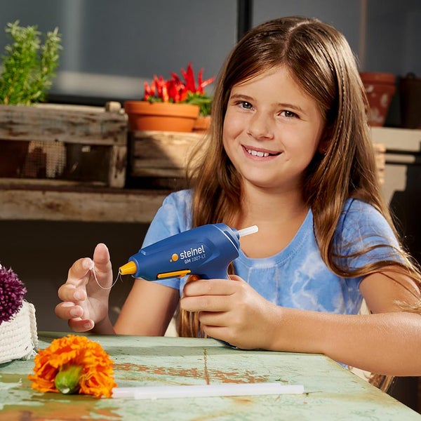 Mädchen nutzt eine Steinel Klebepistole GM 1007-LT zum Basteln an einem Tisch mit Blumen und Klebesticks in einer hellen Umgebung.