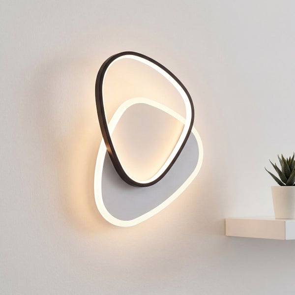 Moderne LED-Wandleuchte mit zwei überlappenden organischen Formen in einer hellen, organisierten Umgebung neben einer Pflanze.