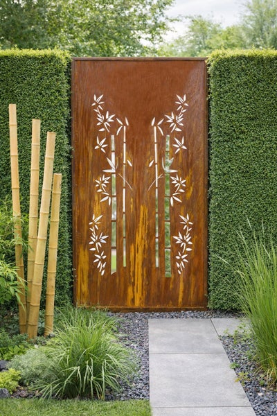 Sichtschutz aus Metall mit Edelrost-Finish und Bambus-Motiv, im Garten zwischen Hecken an einem Steinweg.