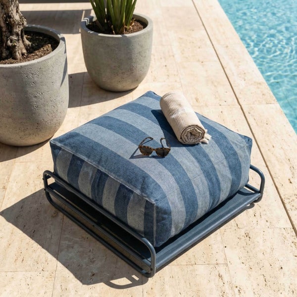 Blau gestreiftes Outdoor-Bodenkissen auf Metallgestell an einem hellen Poolbereich mit Steingefäßen, Handtuch und Sonnenbrille.
