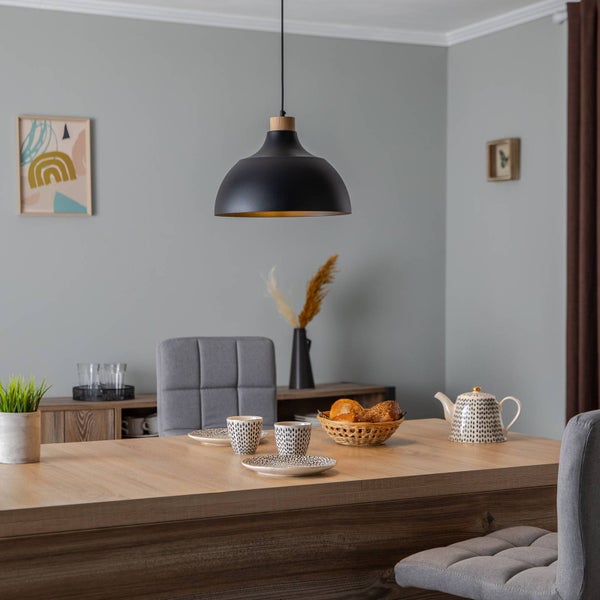 Schwarze Hängeleuchte mit Holzdetail über einem hölzernen Esstisch in einem modern eingerichteten Raum mit grauen Wänden und Stühlen.