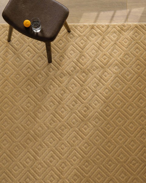 Strukturierter Teppich mit geometrischem Rautenmuster in hellem Braun, auf dem ein kleiner brauner Hocker mit einem Glas Wasser steht.