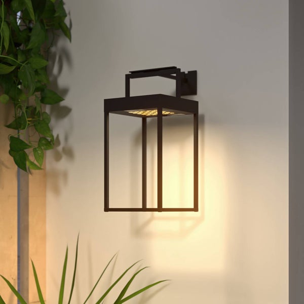 Schwarze LED-Außenwandleuchte in moderner Laternenform an einer Wand neben grünen Pflanzen mit warmem Lichtschein.