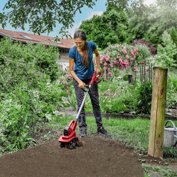 Eine Frau verwendet eine Akku-Bodenhacke zur Bearbeitung von Erde in einem blühenden Garten.