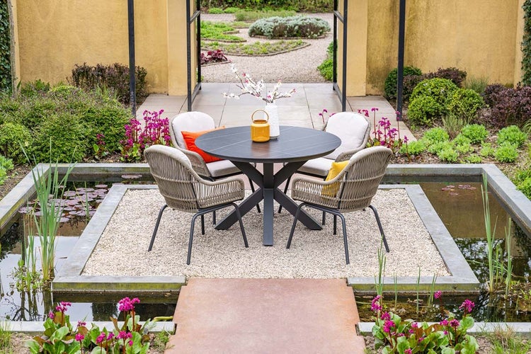 Moderner runder Gartentisch mit vier geflochtenen Gartenstühlen und Kissen auf einer Kiesfläche inmitten eines Gartenteichs.