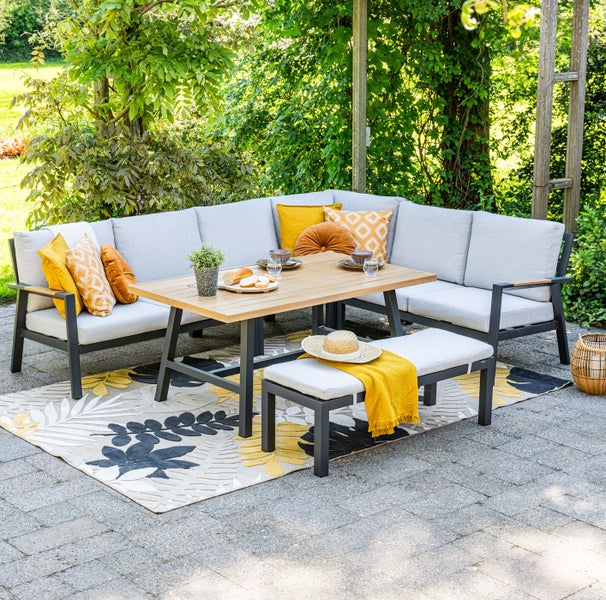 Ecklounge-Set aus Metall mit Holztisch, Bank und hellgrauen Polstern auf einer Terrasse mit Teppich und Pflanzen.