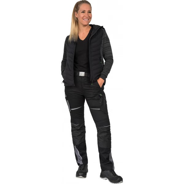 Eine Frau in schwarzer Arbeitskleidung bestehend aus Hybridjacke, Arbeitshose mit Taschen und Sicherheitsschuhen.