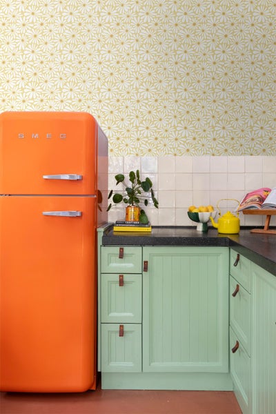 Retro-Küche mit Vliestapete mit Gänseblümchenmuster auf gelbem Grund, Smeg Logo auf orangem Kühlschrank und hellgrünen Küchenschränken.