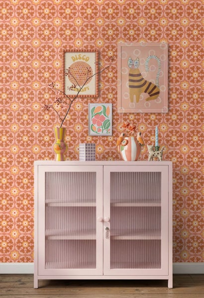 Retro-Zimmer mit gemusterter Blumentapete in Orange und Weiß, einem hellrosa Schrank mit geriffelten Glastüren und verschiedenen Deko-Artikeln.