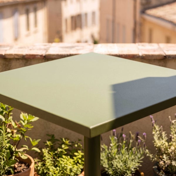 Grüner quadratischer Bistrotisch aus Metall auf einem Balkon mit Pflanzen im Hintergrund.