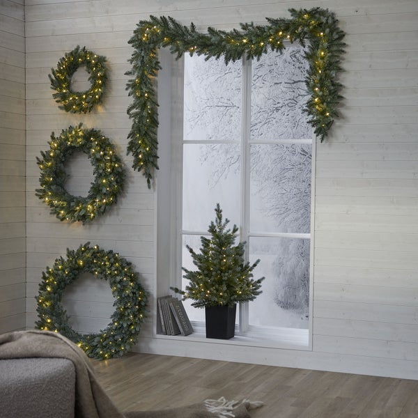 Weihnachtsdekoration mit beleuchteten Kränzen, Girlande und Tannenbaum vor einem Fenster mit winterlicher Aussicht.