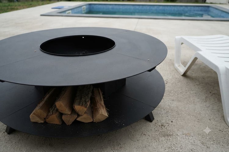 Runde Feuerstelle aus Metall mit Grillplatte und Holzablage auf einer Terrasse vor einem Pool.