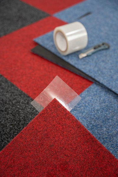 Anwendung von doppelseitigem Klebeband an der Ecke einer Teppichfliese mit einer Rolle Klebeband und einem Cuttermesser im Hintergrund.