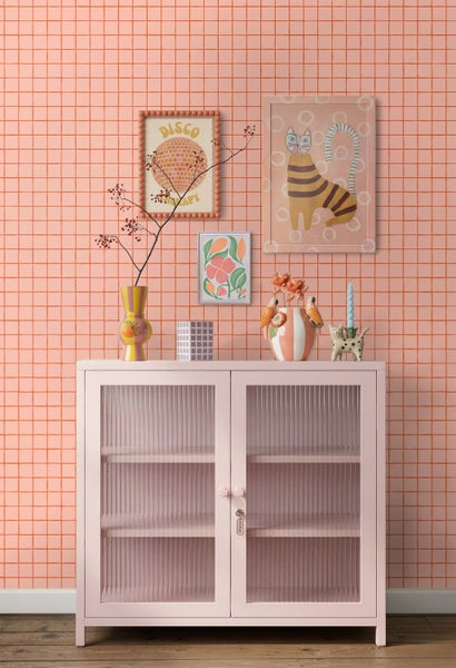 Rosa Vitrinenschrank mit geriffeltem Glas vor einer lachsfarbenen Tapete mit Karomuster und verschiedenen Dekorationsobjekten.