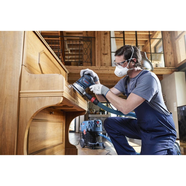 Ein Mann schleift ein Klavier mit einem Exzenterschleifer und Staubsauger in einem hellen Raum.