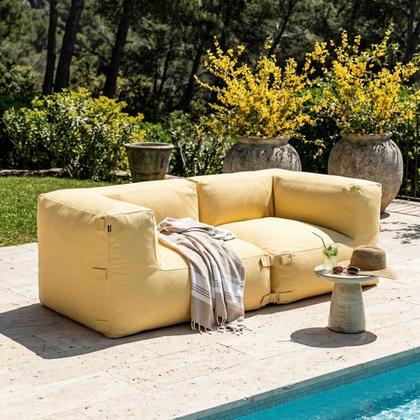 Gelbes modulares Outdoor-Sofa auf einer Steinterrasse am Pool, umgeben von Pflanzen in Steintöpfen und Bäumen im Hintergrund.
