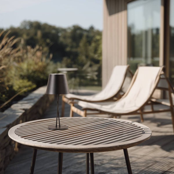 Moderne Tischleuchte auf einem runden Holzlatten-Tisch auf einer hellen Terrasse mit zwei Holz-Liegestühlen und Garten im Hintergrund.