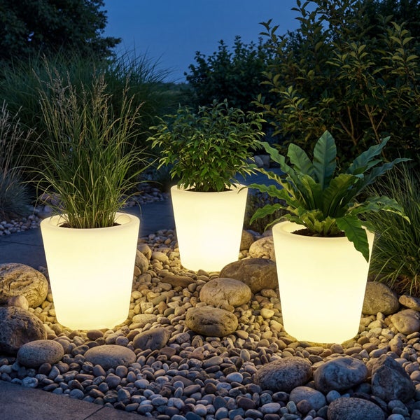 Drei beleuchtete Pflanzkübel aus Kunststoff in einem Steingarten bei Nacht, bepflanzt mit Gräsern und Farnen.