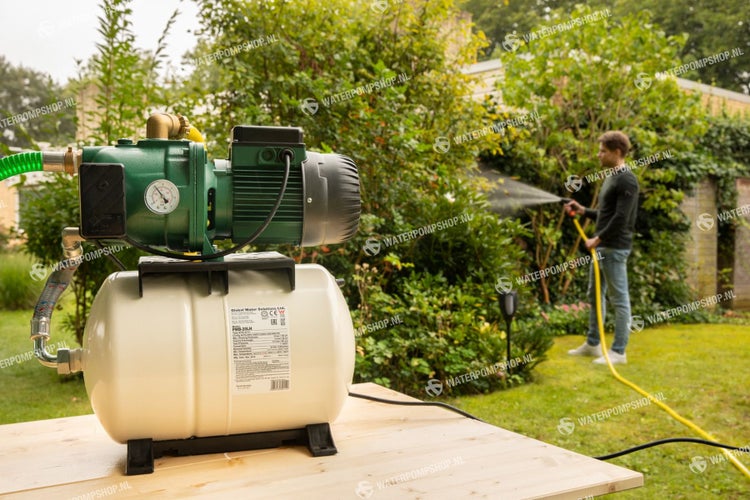 Hauswasserwerk mit Druckkessel auf einem Holztisch, im Hintergrund eine Person mit Gartenschlauch