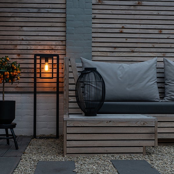 Moderne Außenstehleuchte auf einer Terrasse mit Holzbank, dekorativer Laterne und Pflanze vor einer Holzwand.