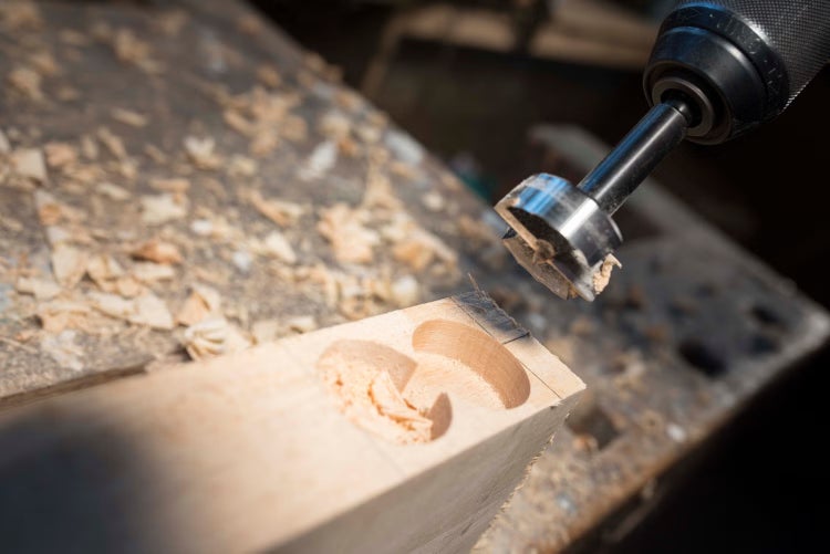 Ein Forstnerbohrer bohrt präzise Sacklöcher in ein Holzstück auf einer Werkbank mit sichtbaren Holzspänen.