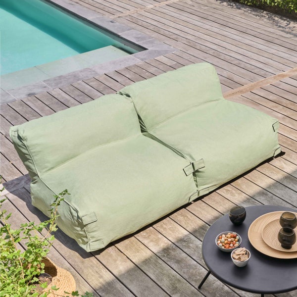 Hellgrünes Outdoor-Loungesofa auf einer Holzterrasse am Pool mit einem runden Beistelltisch in einer hellen Atmosphäre.