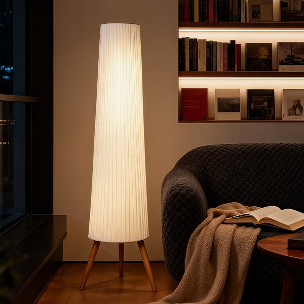 Moderne Stehleuchte mit weißem Plissee-Schirm auf Holz-Stativbeinen in einem Wohnzimmer neben Sofa und beleuchtetem Bücherregal.