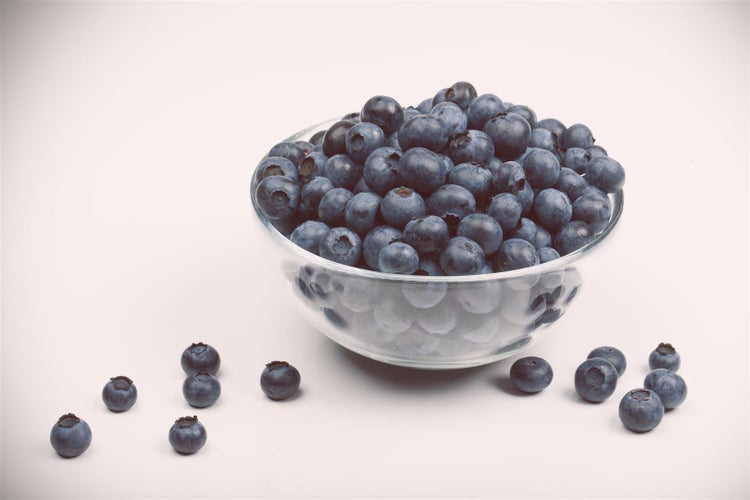 Eine Glasschale gefüllt mit frischen Heidelbeeren sowie einige einzelne Heidelbeeren auf der Oberfläche.
