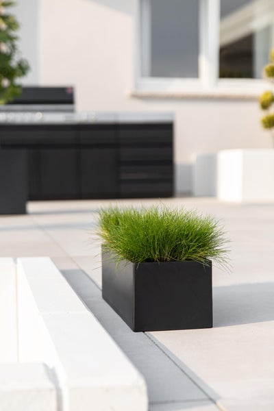 Moderner rechteckiger schwarzer Pflanzkasten mit grünem Gras auf einer hellen Terrasse mit Außenküche im Hintergrund.
