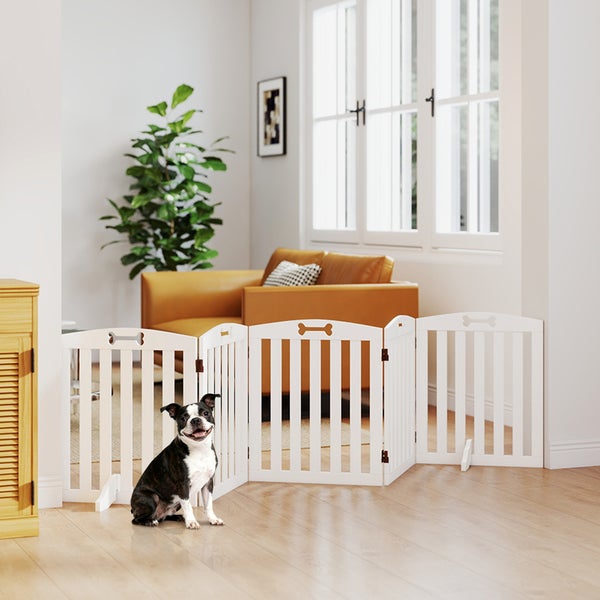 Weißes faltbares Hundeschutzgitter aus Holz mit Knochenmotiven in einem hellen Wohnzimmer vor einem Sofa mit einem kleinen Hund.