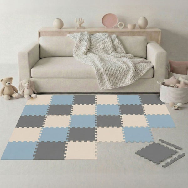 Quadratische Schaumstoff-Puzzlematte in Blau, Grau und Beige vor einem hellen Sofa im Wohnzimmer mit Spielzeug und Dekoration.