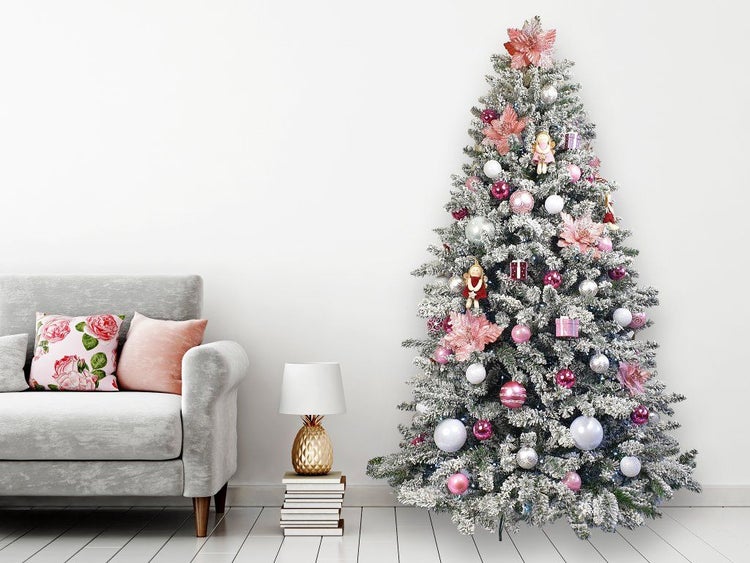 Ein geschmückter, künstlicher Weihnachtsbaum mit Schnee-Effekt neben einem modernen Sofa und einer Tischlampe in einem Wohnzimmer.