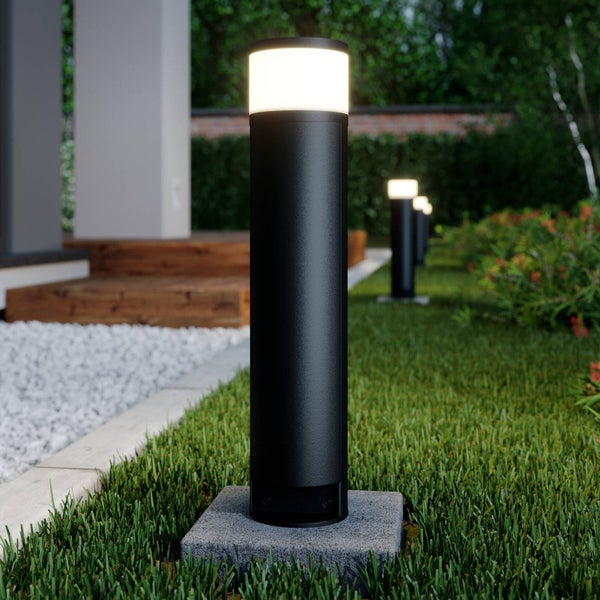 Moderne schwarze Wegeleuchte in Zylinderform auf einer Rasenfläche im Garten neben einem Kiesweg und Holzstufen.