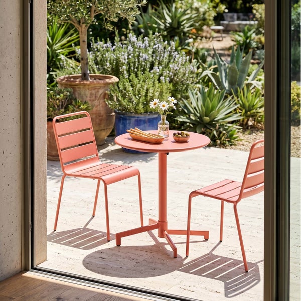Mediterrane Terrassenszene mit rundem Bistrotisch und zwei Metallstühlen in Lachsfarbe auf hellem Steinboden vor einem üppigen Garten.