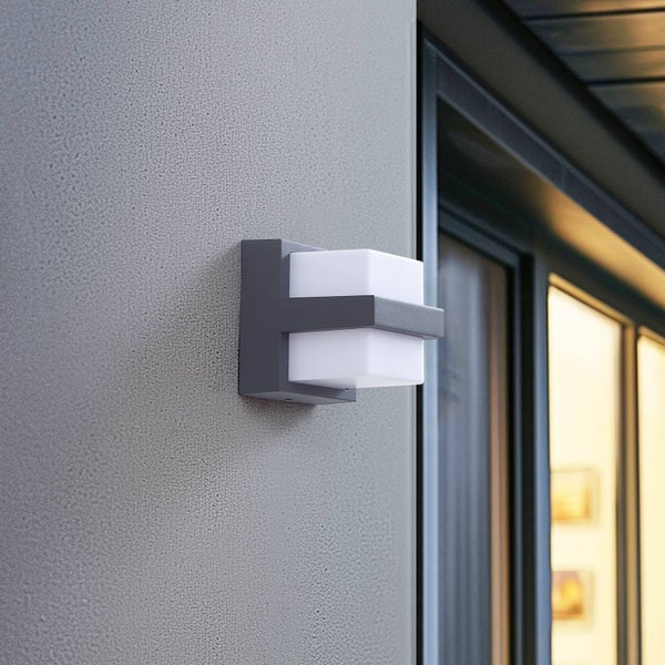 Moderne Außenwandleuchte in Würfelform mit grauem Metallgehäuse und weißem Kunststoff-Diffusor an einer Hauswand neben einem Fenster.