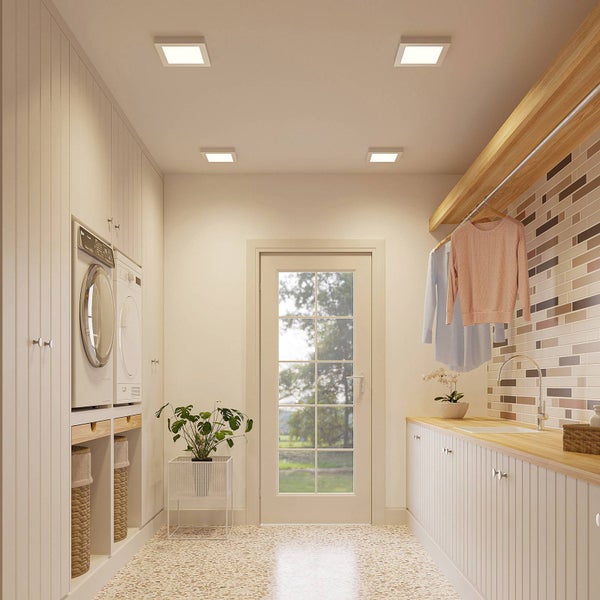 Hell beleuchteter Hauswirtschaftsraum mit vier quadratischen LED Deckenpanels, Waschmaschine, Trockner, Holzarbeitsplatte und Mosaikfliesen.