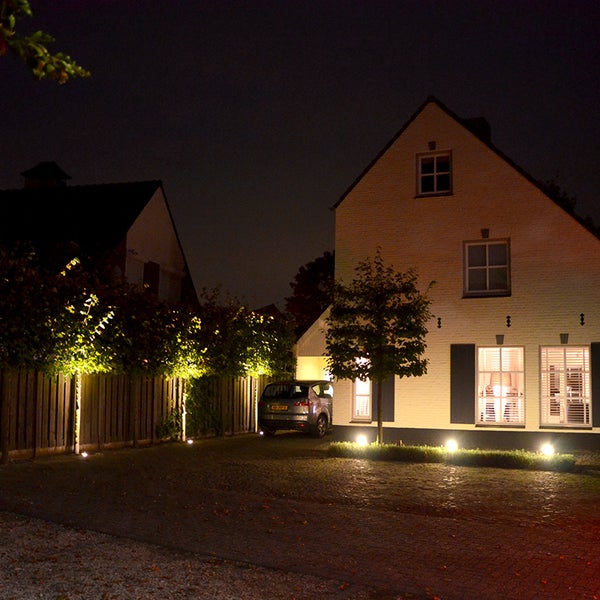 Ein Wohnhaus bei Nacht mit installierter Außenbeleuchtung. Bodenstrahler beleuchten Bäume entlang eines Holzzauns und die Hausfassade.