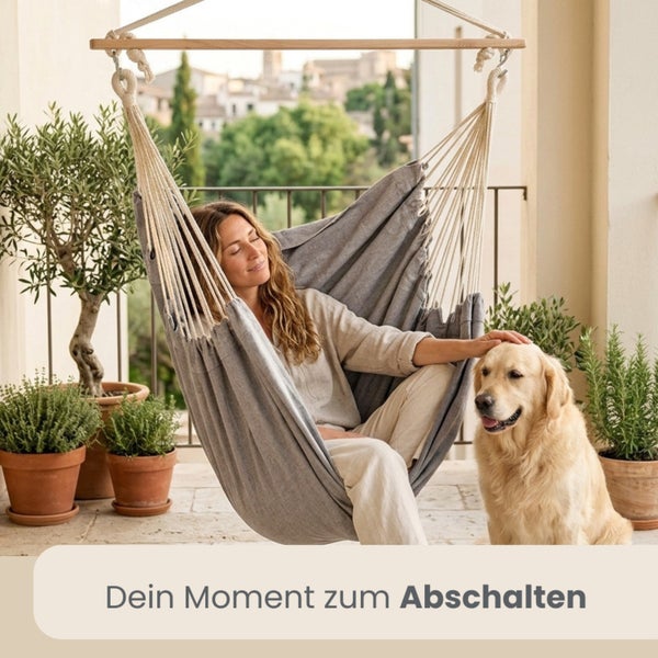 Frau entspannt in einem grauen Hängesessel auf einem hellen Balkon mit einem Hund und Pflanzen in Terrakottatöpfen.