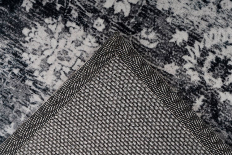 Nahaufnahme einer grau-weißen Teppichecke mit gemustertem Flor in Vintage-Optik und gewebter Rückseite mit verstärkter Kante.