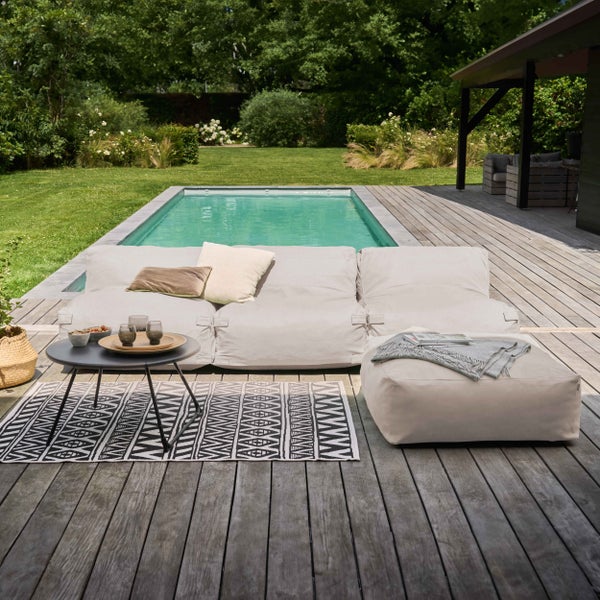 Moderne Gartenlounge aus hellbeigen Sitzelementen auf einer Holzterrasse am Pool, inklusive Beistelltisch und gemustertem Outdoorteppich.