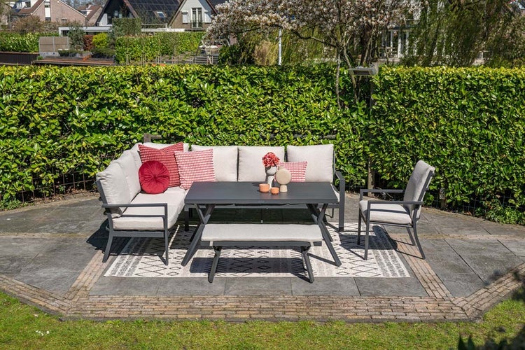 Modernes Gartenmöbel-Set aus grauem Aluminium mit Ecksofa, Esstisch, Bank und Sessel auf einer Steinterrasse vor einer grünen Hecke.