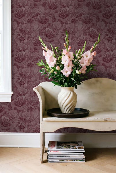 Eine Wand mit dunkelroter Blütentapete, davor eine weiße antike Bank mit einer Vase voll rosa Gladiolen und Zeitschriften auf dem Boden.